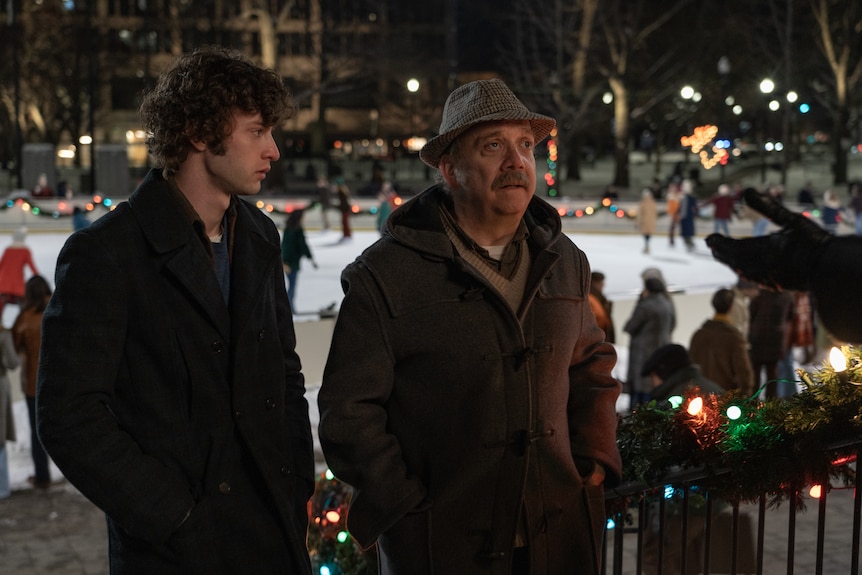 A film still of Dominic Sessa and Paul Giamatti standing outside an ice-skating rink, looking miserable.