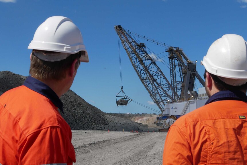 Two men wearing bright orange shirts and hard hats with backs to the camera looking at a dragline