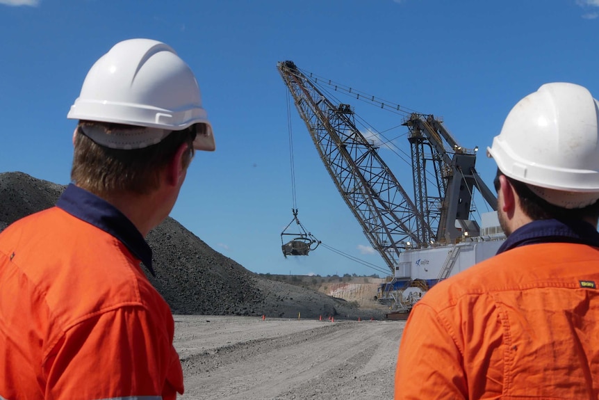 Two men wearing bright orange shirts and hard hats with backs to the camera looking at a dragline