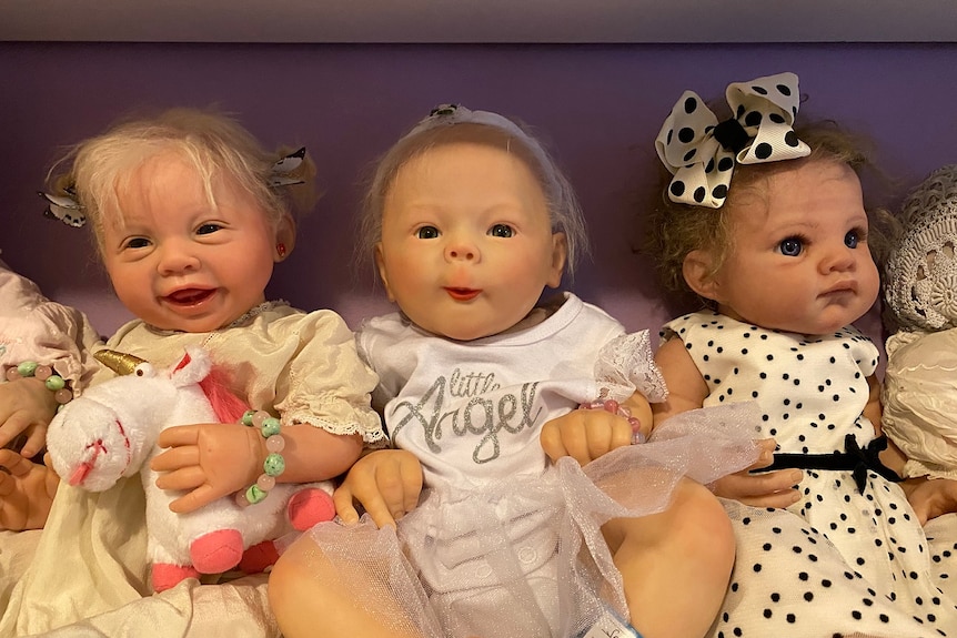 Three lifelike dolls sit together on a shelf inside a doll museum.