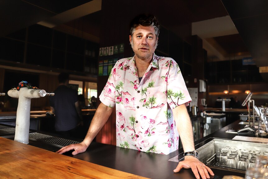 Man wearing a floral shirt stands with his hands atop a wooden bar with pub in the background 