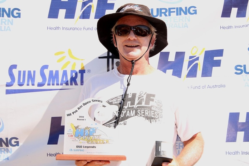 A man with a hat and sunglasses on standing in front of a backdrop holding two surfing trophies. 