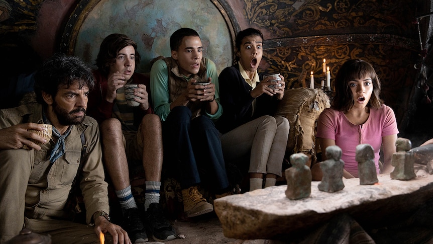 A man and four teenagers sit with shocked expressions while looking at four artefacts sitting on table.