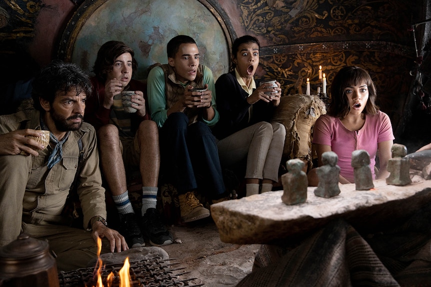 A man and four teenagers sit with shocked expressions while looking at four artefacts sitting on table.