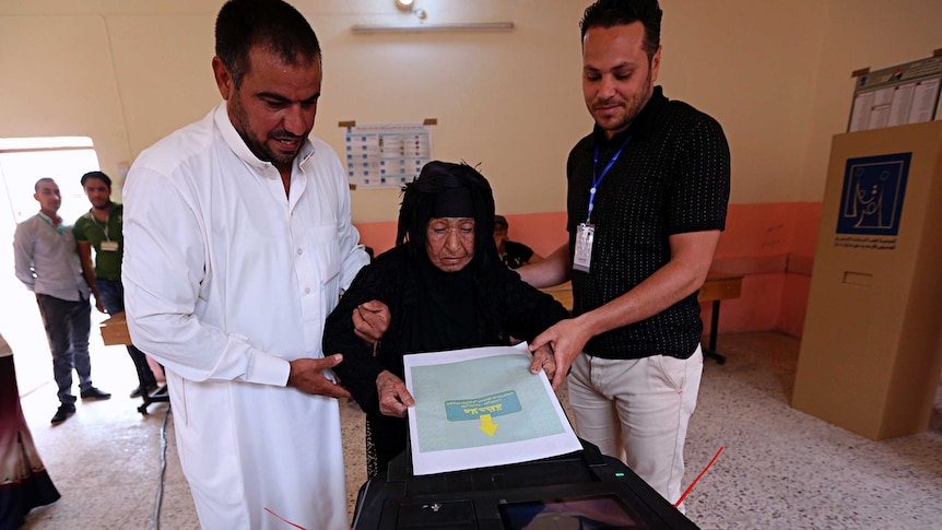 An Iraqi elderly woman casts her ballot, assisted by two men.
