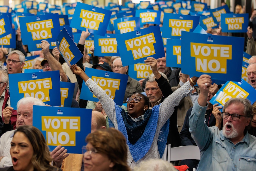 A huge crowd yells and holds up blue signs that say 'Vote no'.