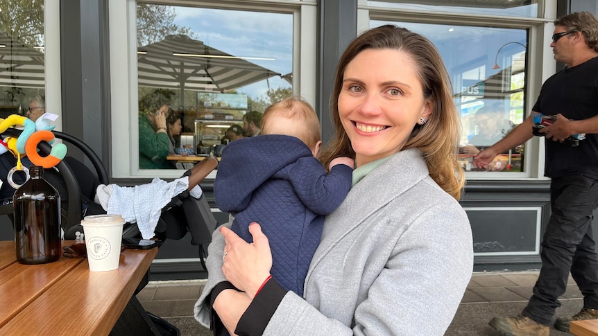 woman in grey coat smiles at camera, while cradling baby against her shoulder and sitting at a table outside busy café 