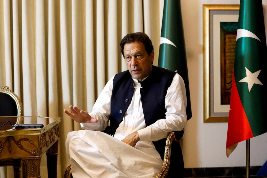 An older Pakistani man sits at a desk in front of the Pakistani flag.