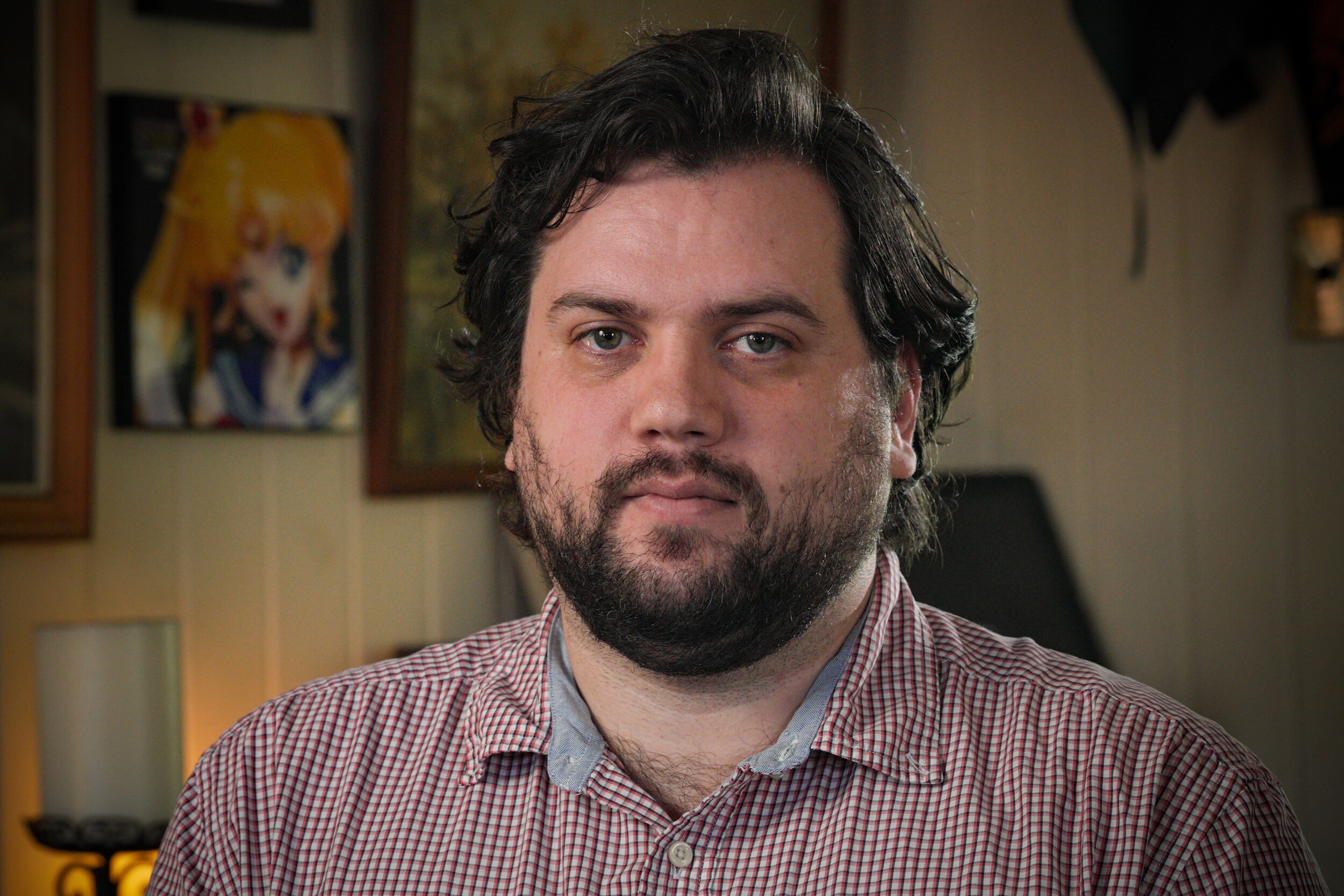 A young man with a beard poses for a portrait inside, looking into camera with a serious facial expression.
