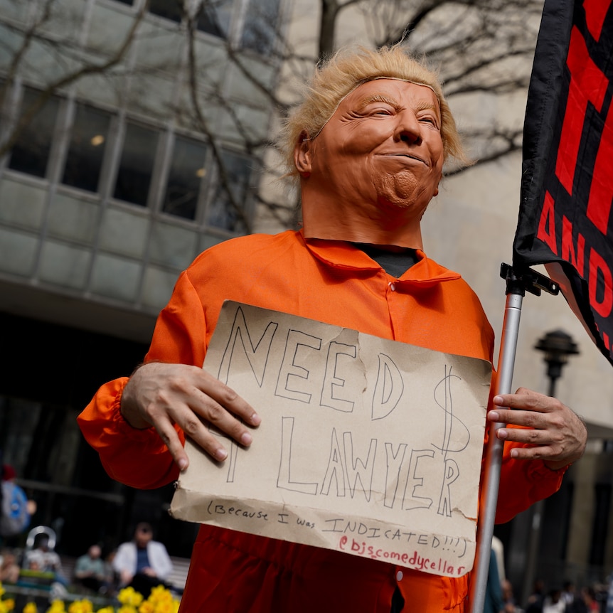 A person in an orange jumpsuit wearing a Donald Trump mask holds a sign saying NEED A LAWYER
