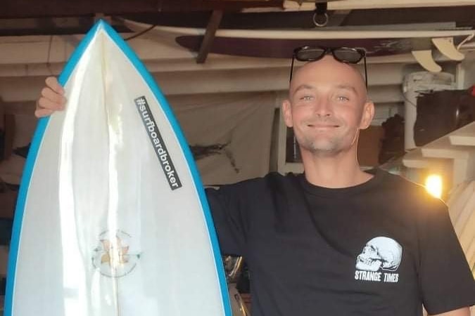 A man stands smiling, holding a surfboard inside a shed.