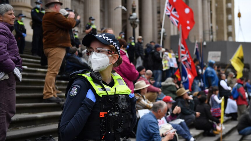 A masked female police officer stands on the steps of the Parliament of Victoria as protesters gather behind her.