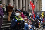 A masked female police officer stands on the steps of the Parliament of Victoria as protesters gather behind her.