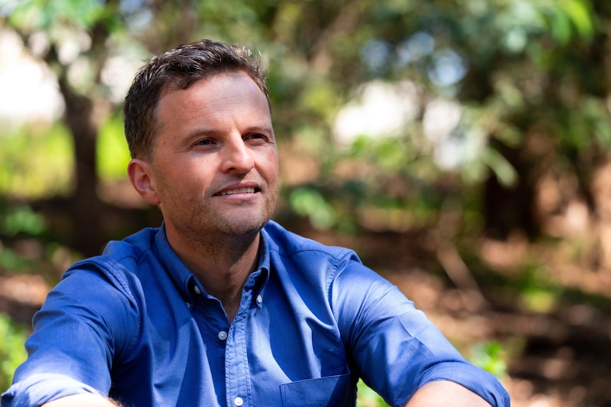 A man in a blue short looks away from the camera among a leafy background. 