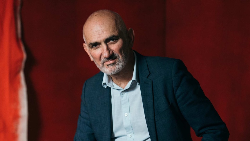 Australian singer-songwriter Paul Kelly poses in front of a red backdrop.