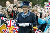 queen elizabeth II wearing a dark blue coat and hat stands in front of a crow of children holding British flags