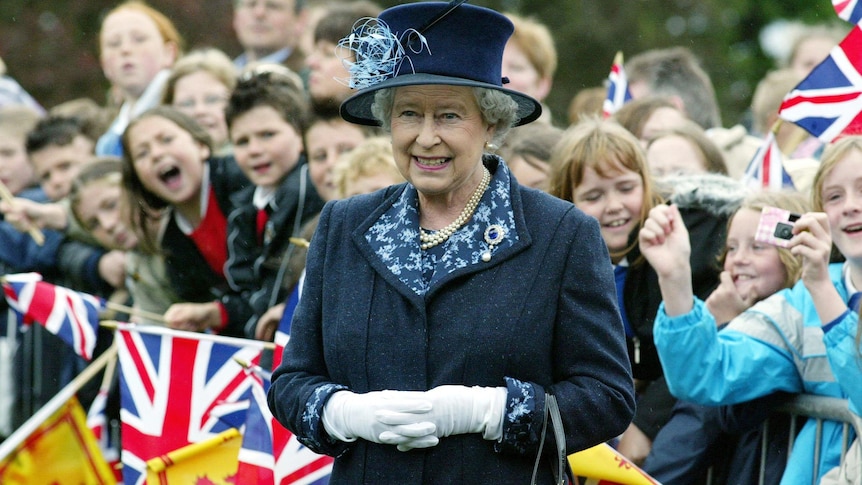 queen elizabeth II wearing a dark blue coat and hat stands in front of a crow of children holding British flags