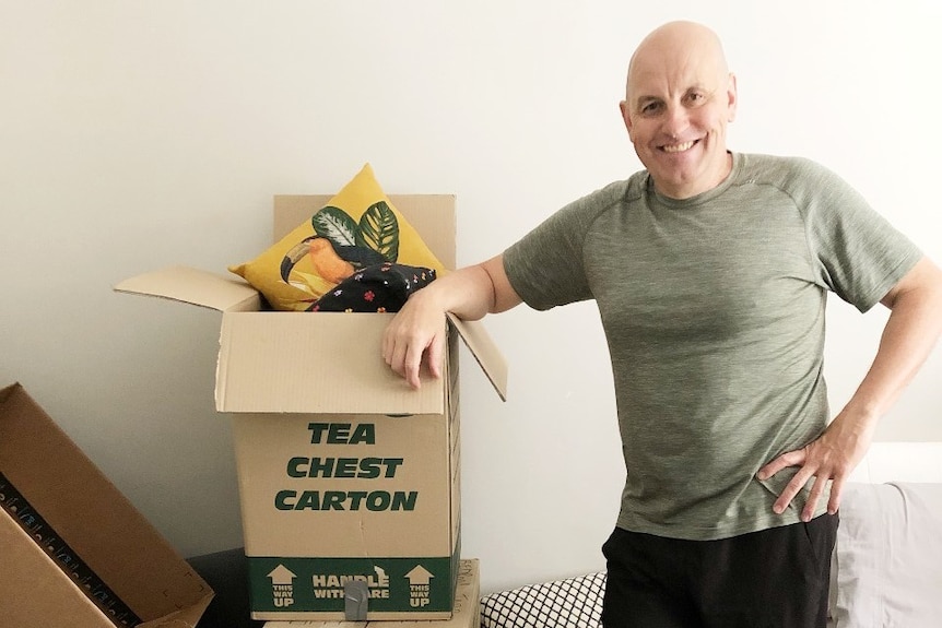 A balding man stands next to packing boxes