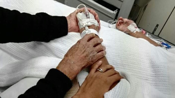 Two people wearing black and grey jumpers hold a girl's hand on a hospital bed.