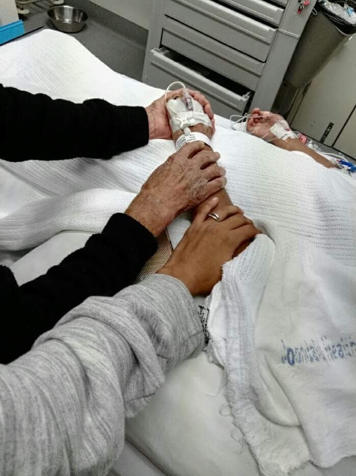 Two people wearing black and grey jumpers hold a girl's hand on a hospital bed.
