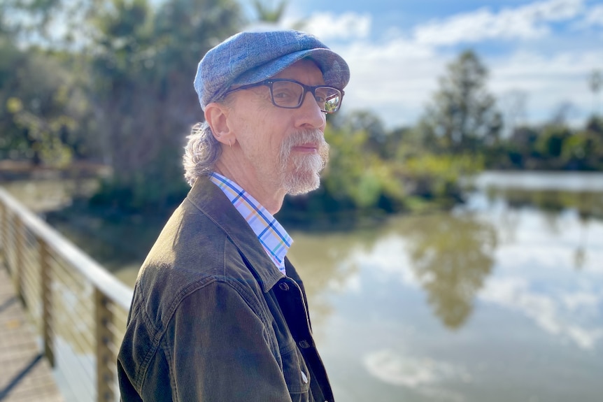 a man with a beard wearing glasses and a hat. there is a lake behind him.