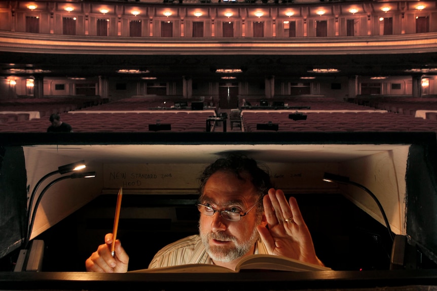 A man in an opera prompter box on stage, with a threatre behind.