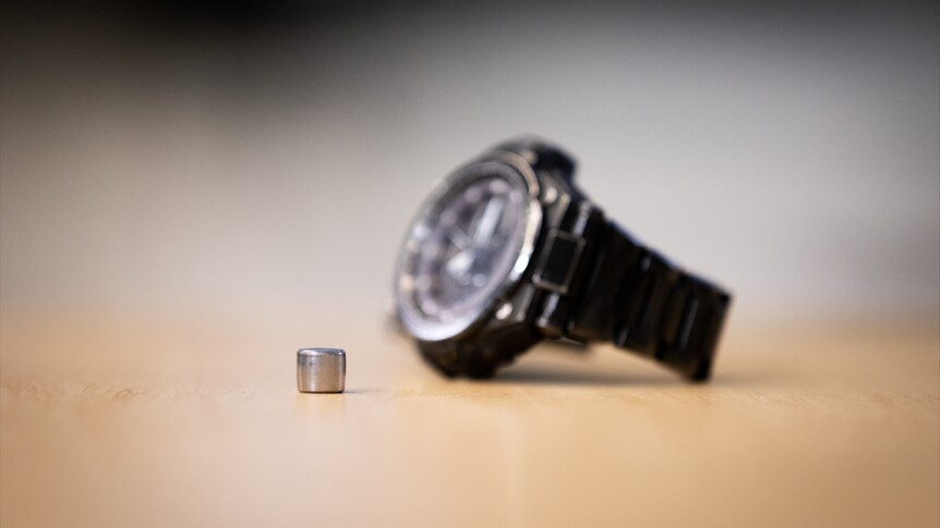 A tiny battery-like metal object on a table next to a wristwatch.
