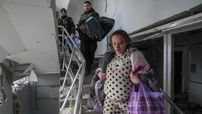 A pregnant woman with blood on her face and carrying a number of bags walks down a set of damaged stairs. Two men follow behind.