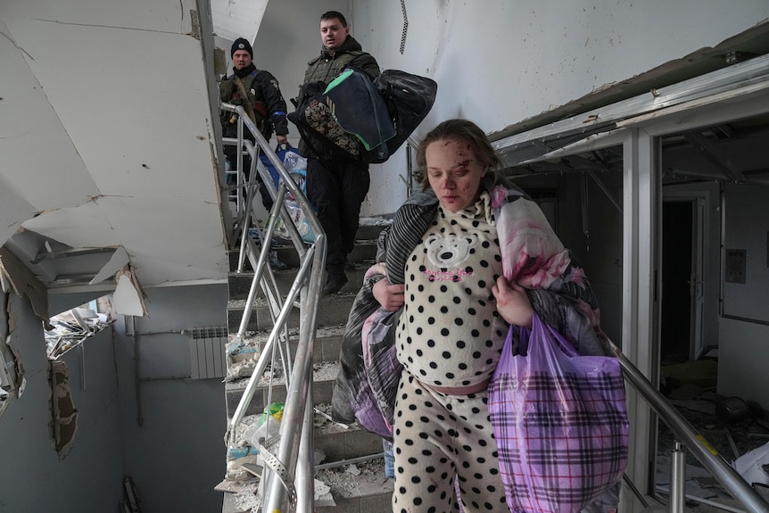 A pregnant woman with blood on her face and carrying a number of bags walks down a set of damaged stairs. Two men follow behind.