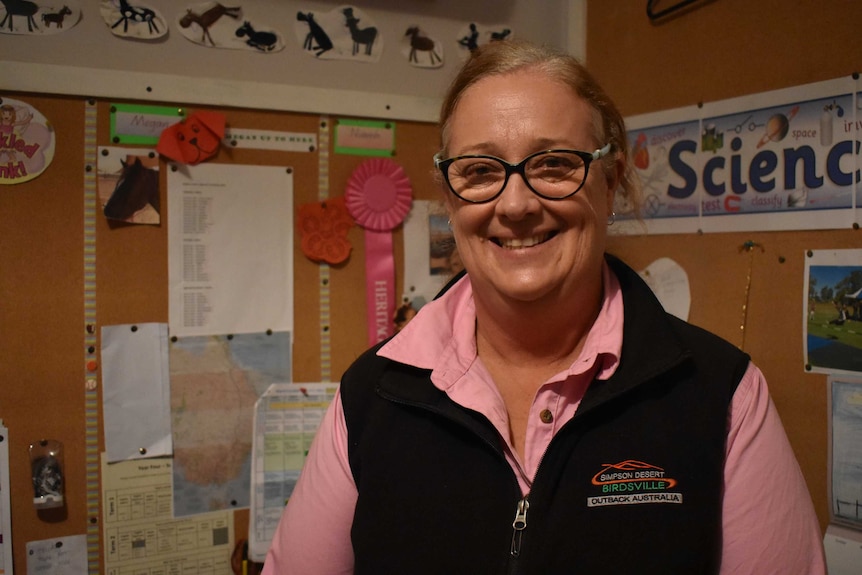 A woman smiles wide, wearing black round glasses with teaching paraphernalia on the walls in the background.