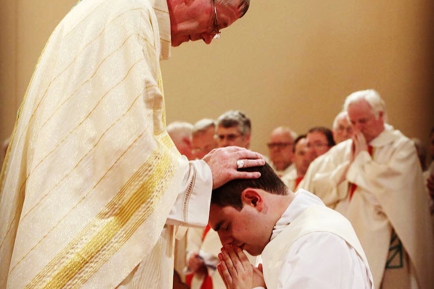 An older priest places his hands on a kneeling David's head as he clasps his hands together in prayer