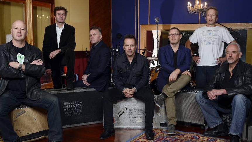 Seven members of Hunters and Collectors sit on top of amplifiers and roadcases in a music studio