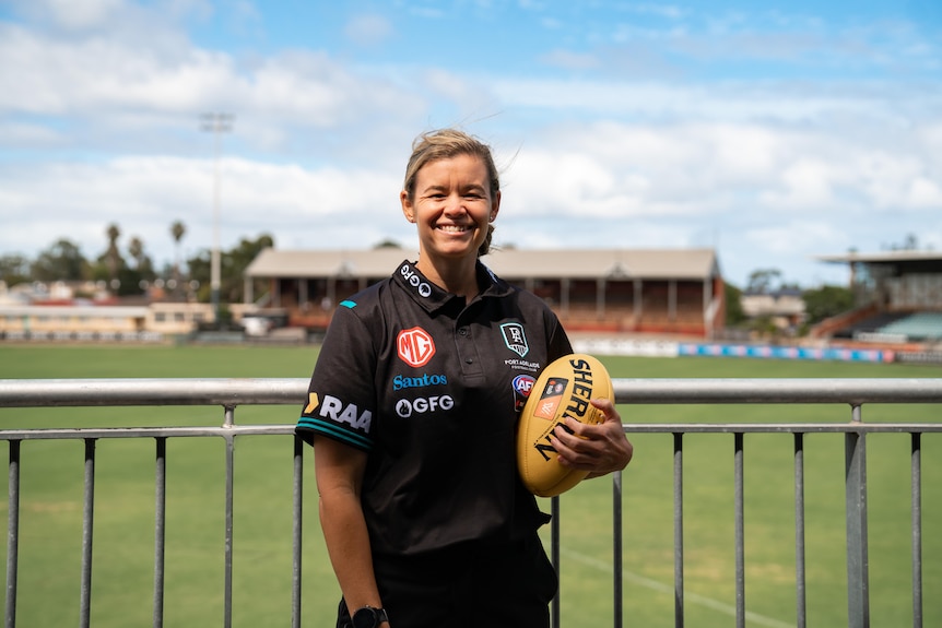 A woman in Port Adelaide football club polo shirt holding a football next to the oval