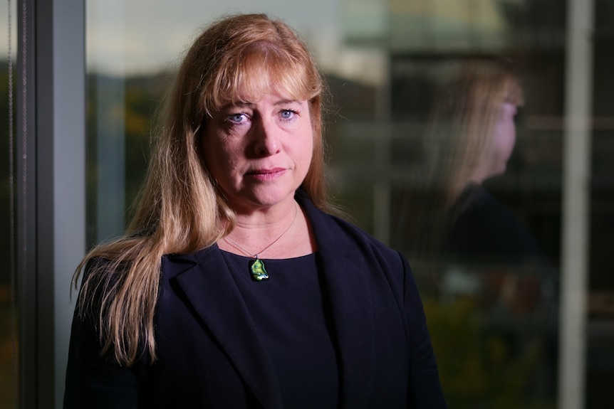 A woman stands in an office and looks at the camera with a serious expression on her face.