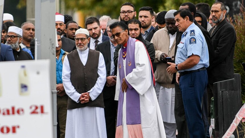 A group of solemn people, some wearing religious attire, stand in a group outside a mosque.