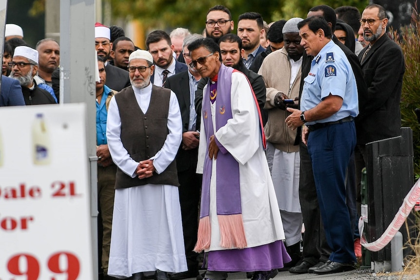 A group of solemn people, some wearing religious attire, stand in a group outside a mosque.