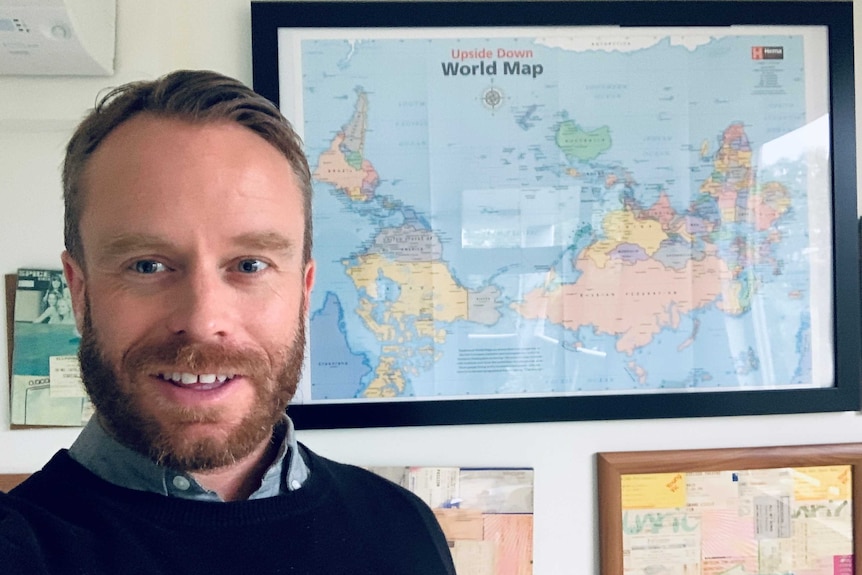A photo of a man standing in front of a framed map of the world