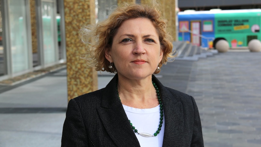 A woman with curly hair in a suit stands in an outdoor square.