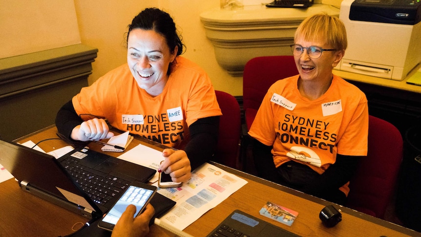 Two female volunteers help an attendee across the desk with their phone.