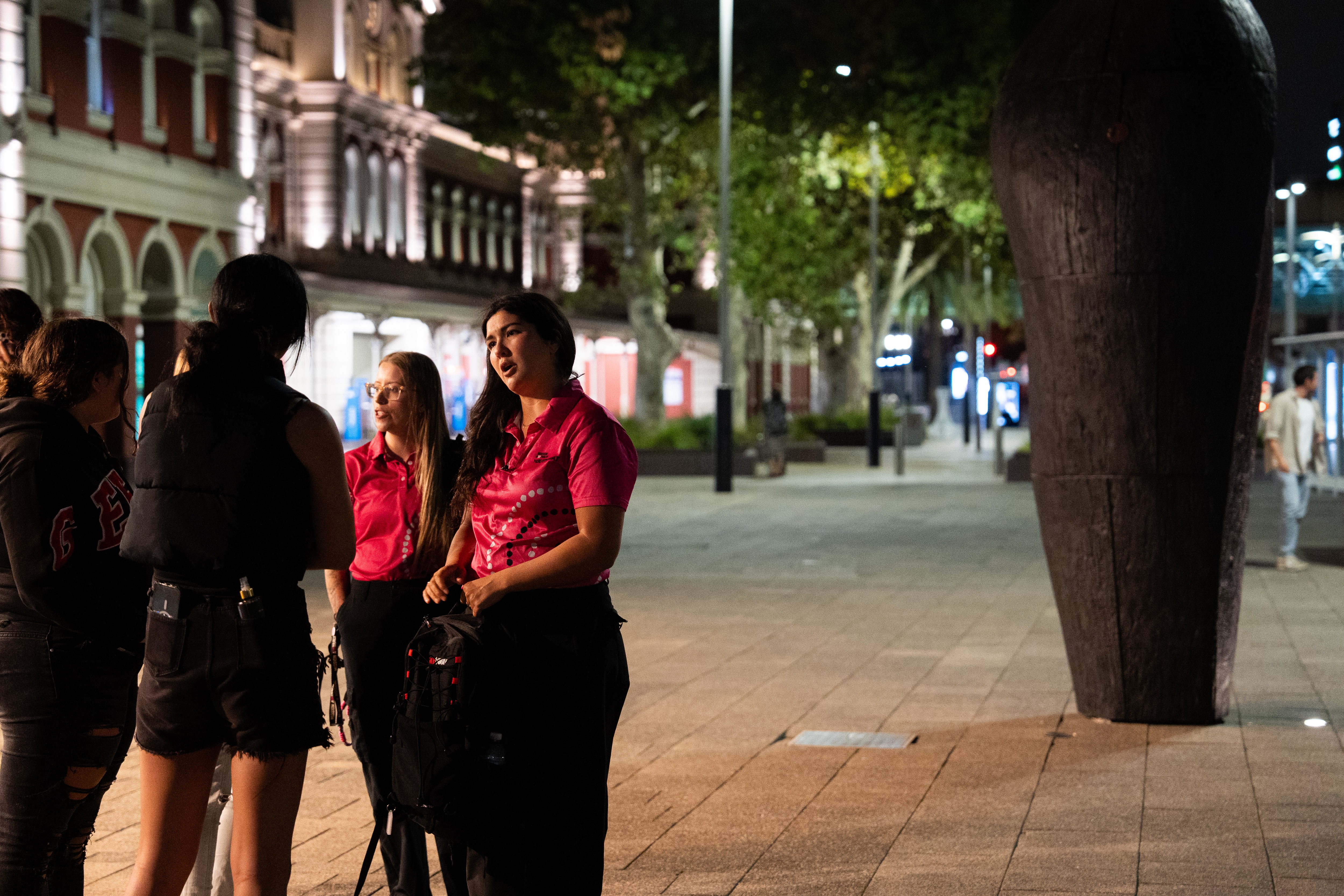 two female youth workers in pink shirts talk to a froup of teenage girls outside Perth train station.