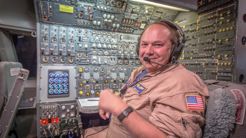 A man wearing NASA jumpsuit smiles while sitting in front of an array of dials.