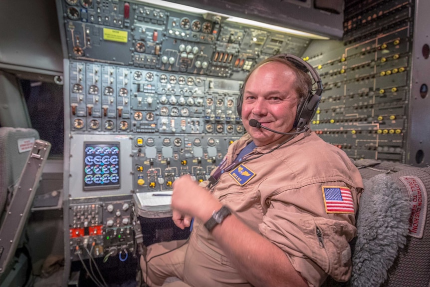 A man wearing NASA jumpsuit smiles while sitting in front of an array of dials.