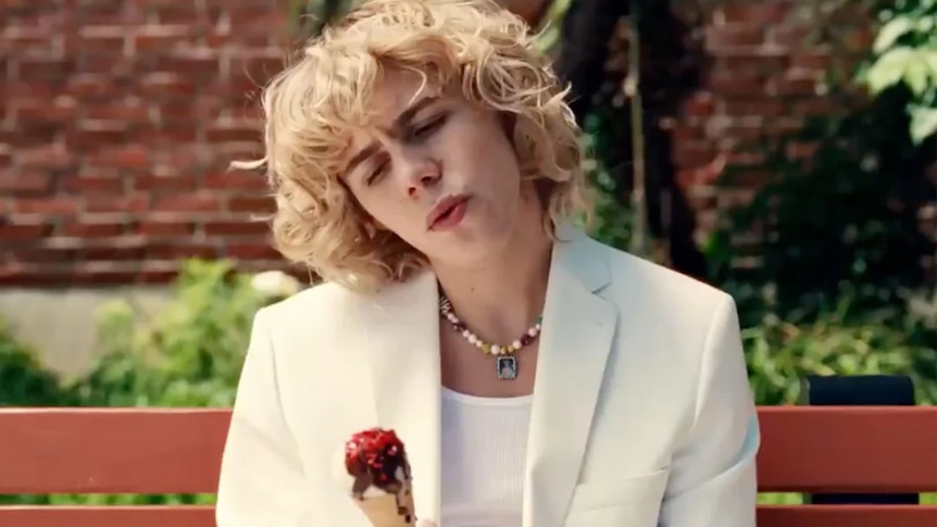 Male with blonde curly hair sits on a red bench, wearing white suit and shirt, holding an ice cream cone