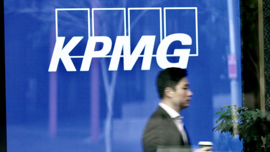 A man wearing a suit carrying a cup of coffee walks past a KPMG logo.
