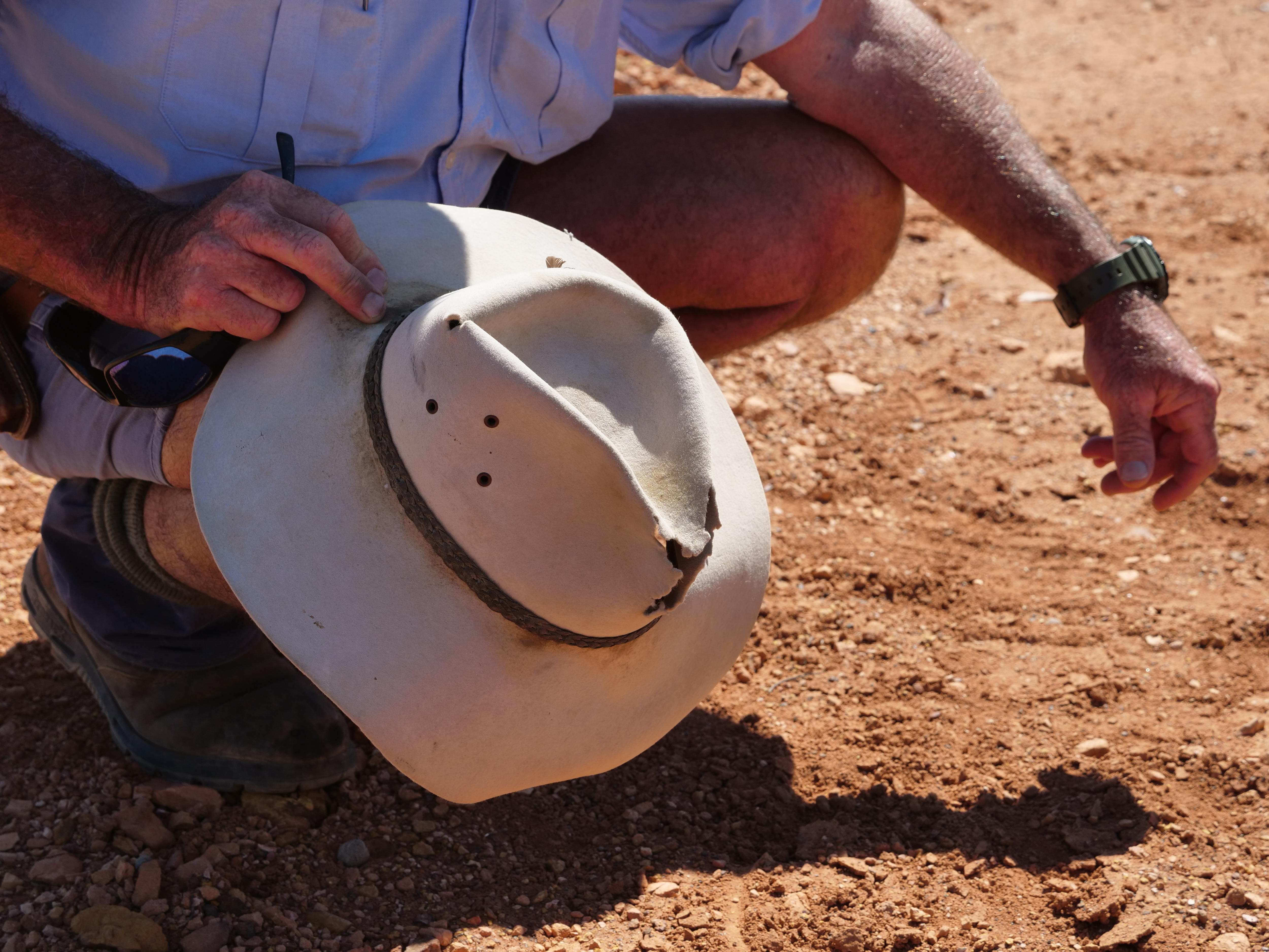 Un hombre se agacha sobre tierra roja y sostiene un sombrero de vaquero en la rodilla. 