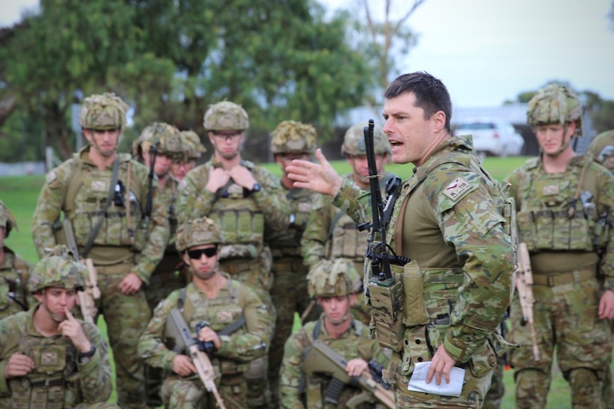 An Australian Army major gives orders to his soldiers who are huddled around him in full combat gear in an open outdoor space.