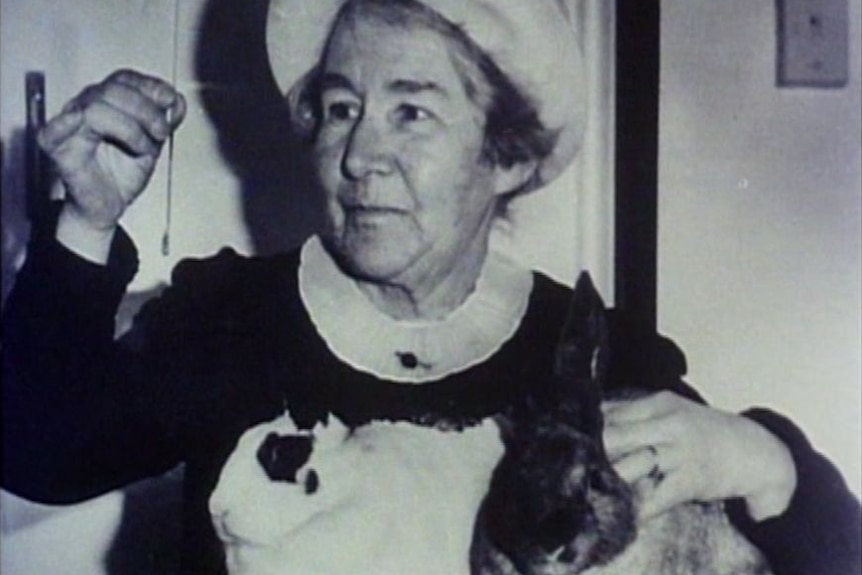 A black and white image of a middle-aged woman holding a rabbit and a needle.