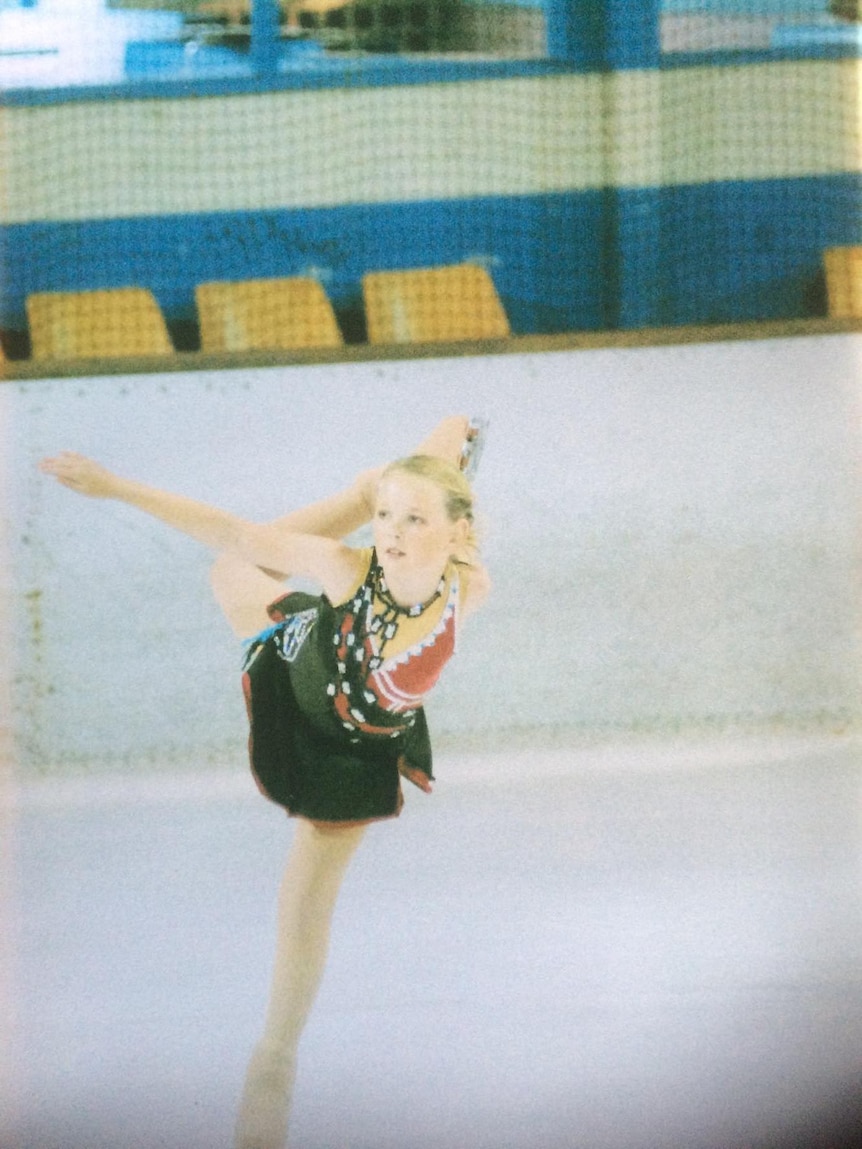 A young girl skates on ice in a black leotard and skirt, balancing on one leg with the other held up behind her.