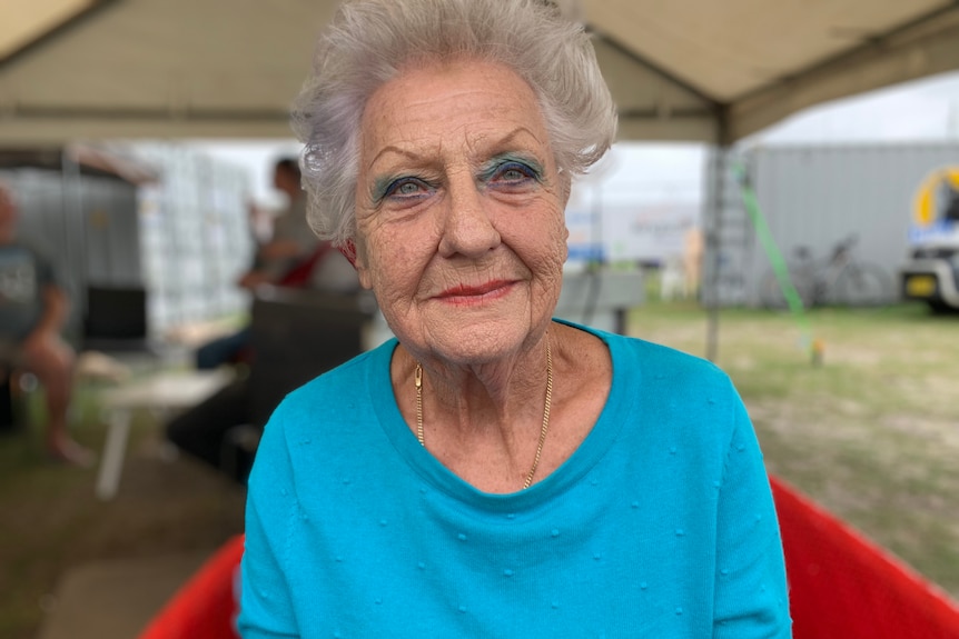 An older woman with grey hair and a blue T-shirt looks into the camera.