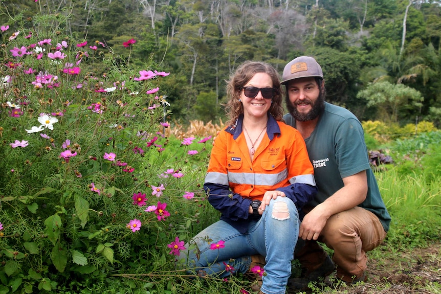 A man and a woman pose in their veggie patch with pink and white flowers to their left